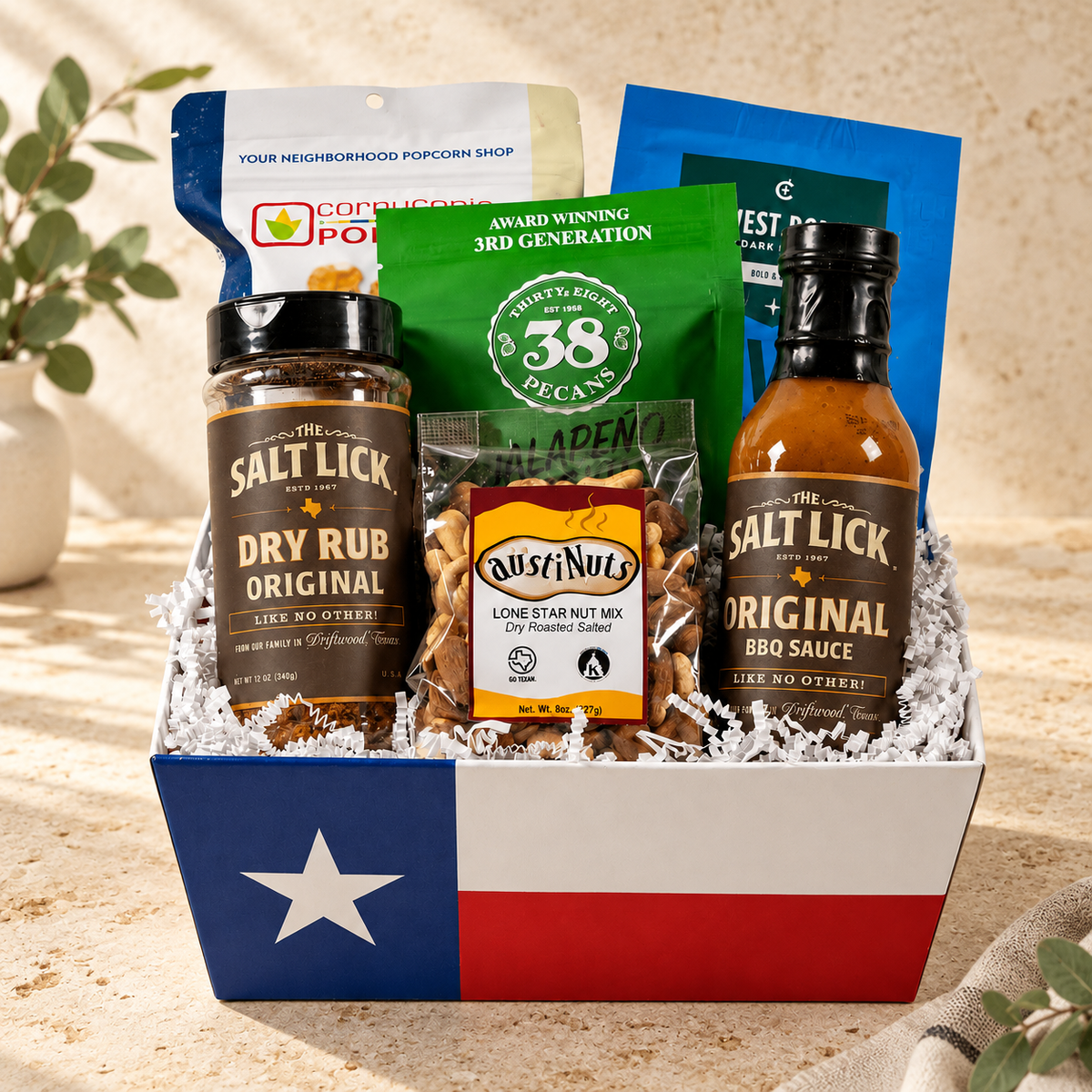 A gift basket styled with Texas flag colors (blue, white, and red) containing a bottle of Salt Lick Dry Rub Original, a bottle of Salt Lick Original BBQ Sauce, a bag of Rusty Nuts Lone Star Nut Mix, and other snacks on a sandy background.